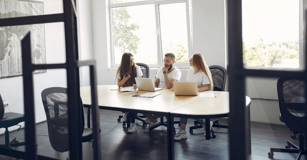 Drei Personen sitzen mit Laptops an einem Konferenztisch und diskutieren in einem hellen, dank der Büroreinigung Graz tadellos gepflegten Büroambiente.