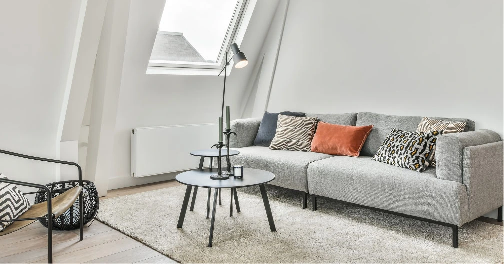 Modernes Wohnzimmer in Graz mit einem grauen Sofa, verschiedenen Kissen, zwei runden schwarzen Couchtischen, einer Stehlampe und einem Sessel unter einem Oberlichtfenster. Der beige Teppich auf dem Holzboden unterstreicht den makellosen Look des Raums nach einer gründlichen Grundreinigung.