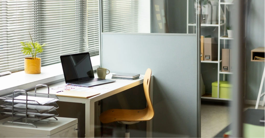 Ein modernes Büro in Graz verfügt über einen Schreibtisch mit Laptop, Stuhl, Tasse, Notizbüchern und einer kleinen Topfpflanze am Fenster mit Jalousien. Sorgen Sie mit erstklassigen Büroreinigungsdiensten in der Gegend dafür, dass Ihr Arbeitsplatz makellos bleibt.